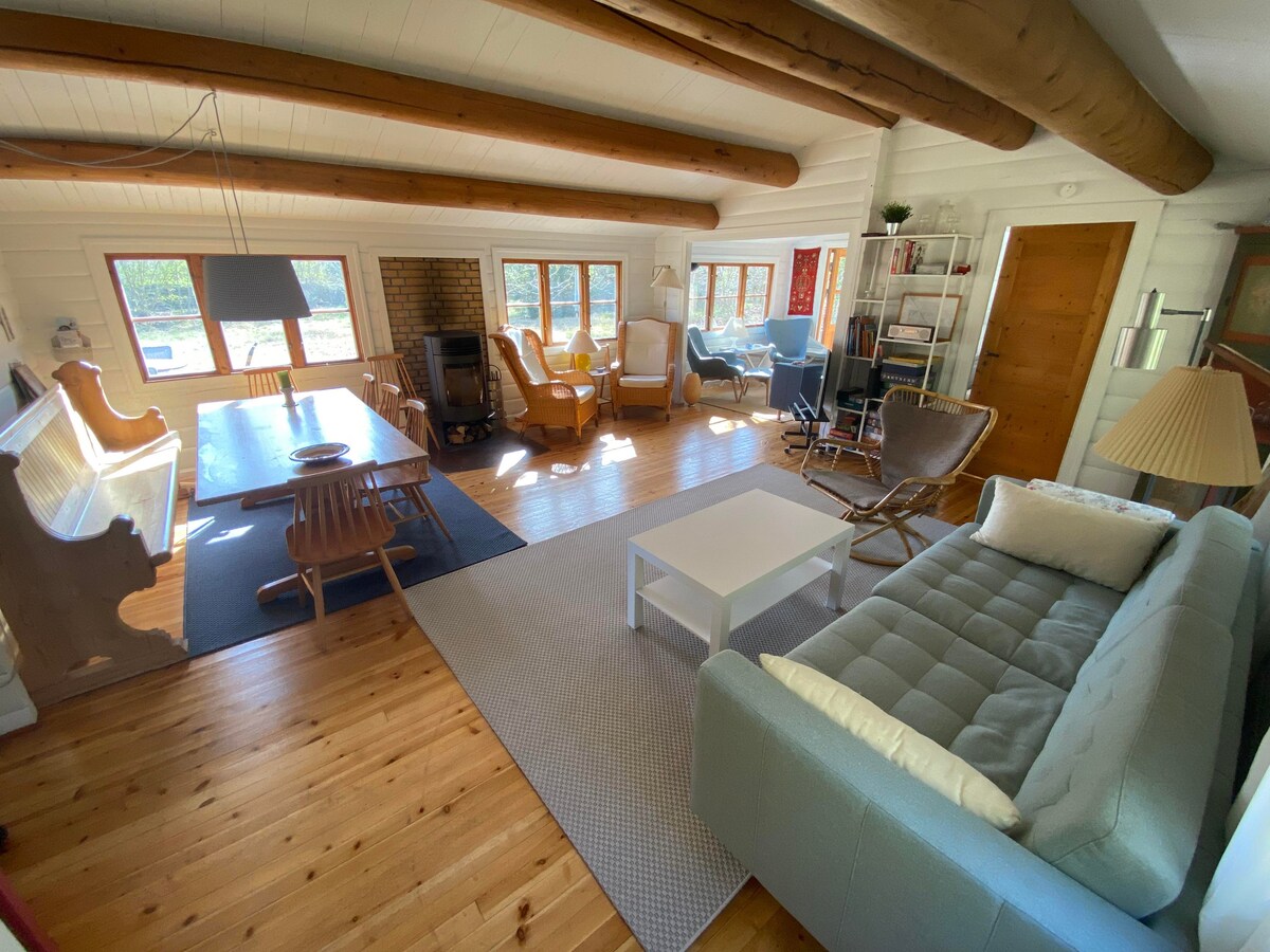 The living area showcases a spacious layout with natural light streaming through multiple windows. A dining table and chairs are positioned near the center, while various seating options, including a comfortable sofa and lounge chairs, invite relaxation. Wooden beams adorn the ceiling, adding a rustic touch.