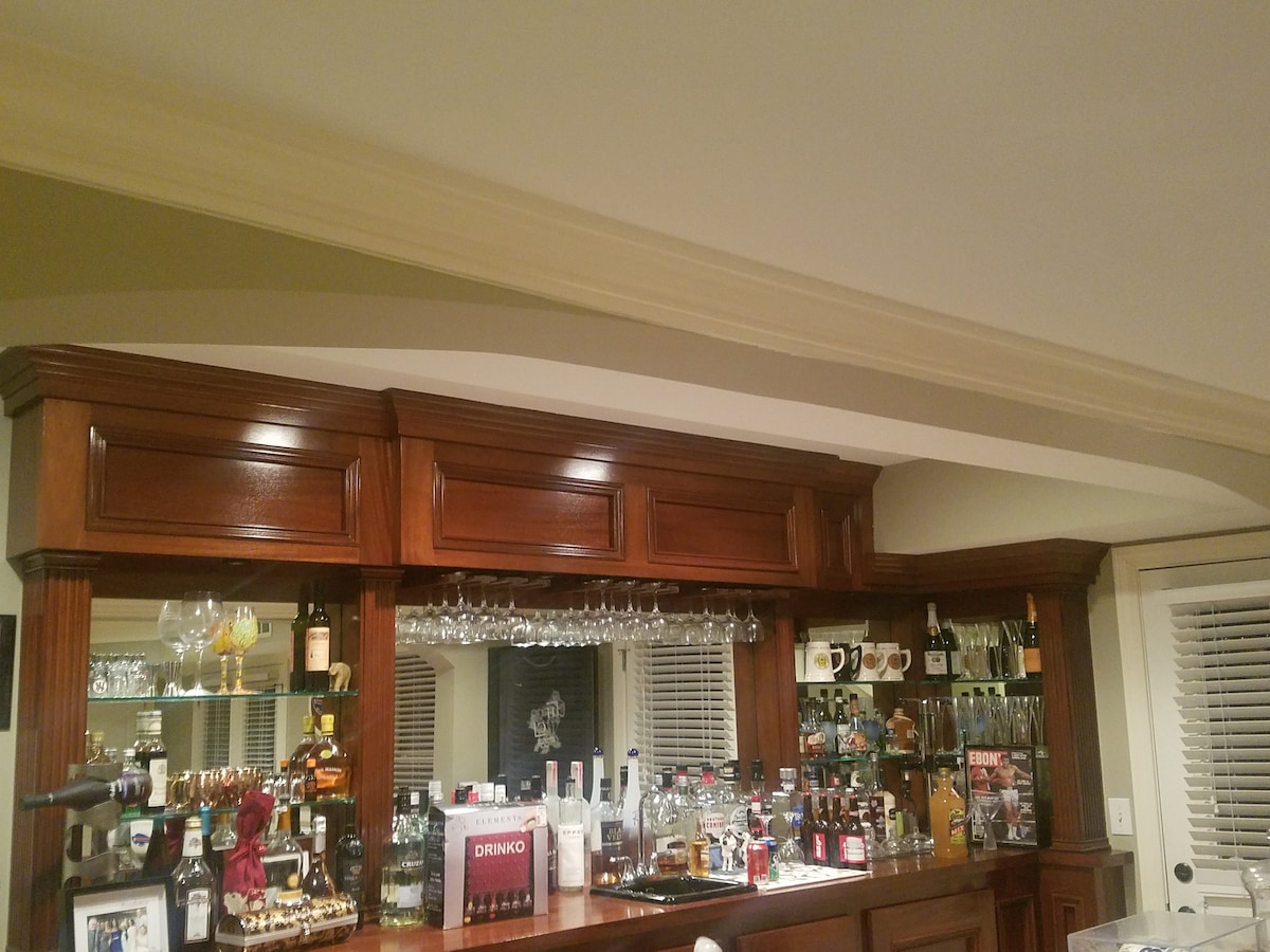 A polished wooden bar showcases an array of glassware and spirits, with various liquor bottles displayed behind. The elegant design features cabinetry and a countertop, enhancing the inviting setting. Soft lighting reflects subtly from the surfaces, adding to the overall ambience.