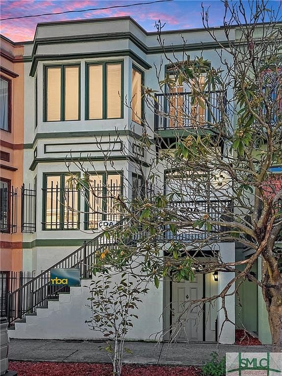 A charming multi-level building is presented, featuring a warm color palette and intricate architectural details. The exterior includes green accents and large windows framed by elegant black railings. Surrounding greenery adds a touch of nature, enhancing the inviting appearance of the entrance.