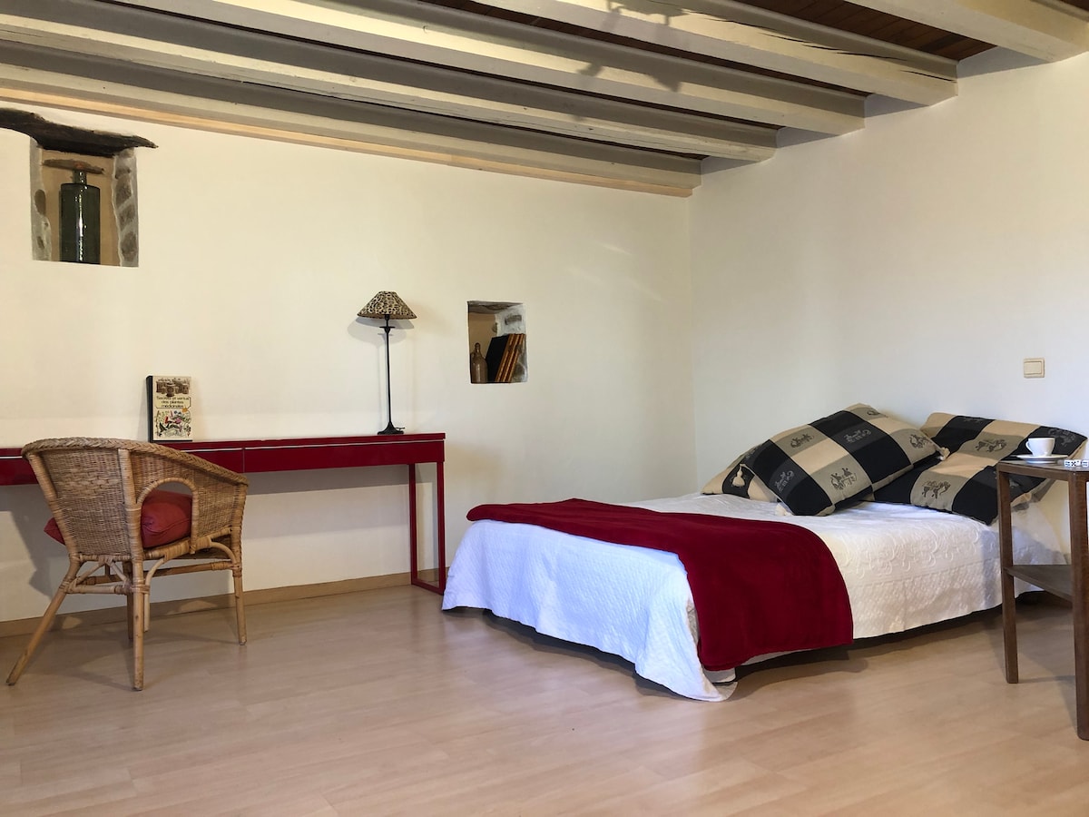A bright bedroom is presented, featuring a spacious bed dressed in white linens with decorative pillows. A desk and a chair are positioned near a wall, while natural light filters through a small window. The wooden ceiling beams add warmth to the room.