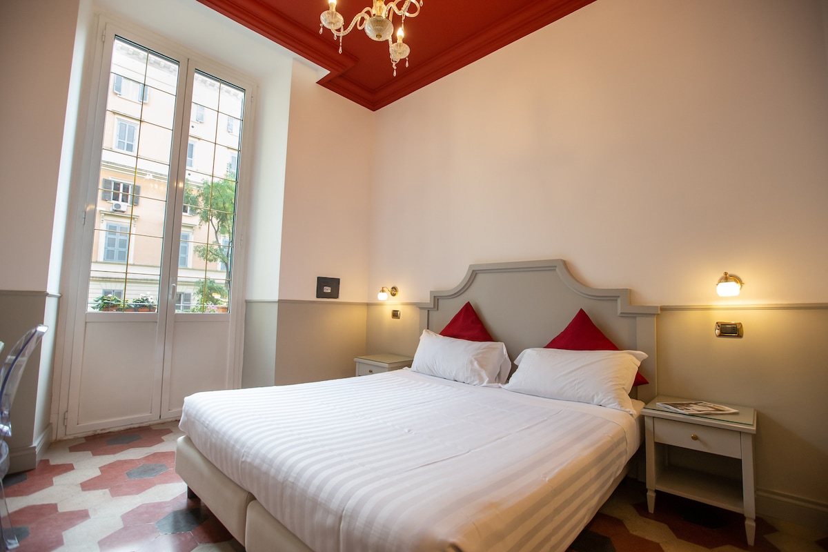 A well-lit bedroom features a spacious bed adorned with white linens and red accent pillows. Two bedside tables with lamps are present, and large windows allow natural light to fill the space while offering a view of the outdoors.