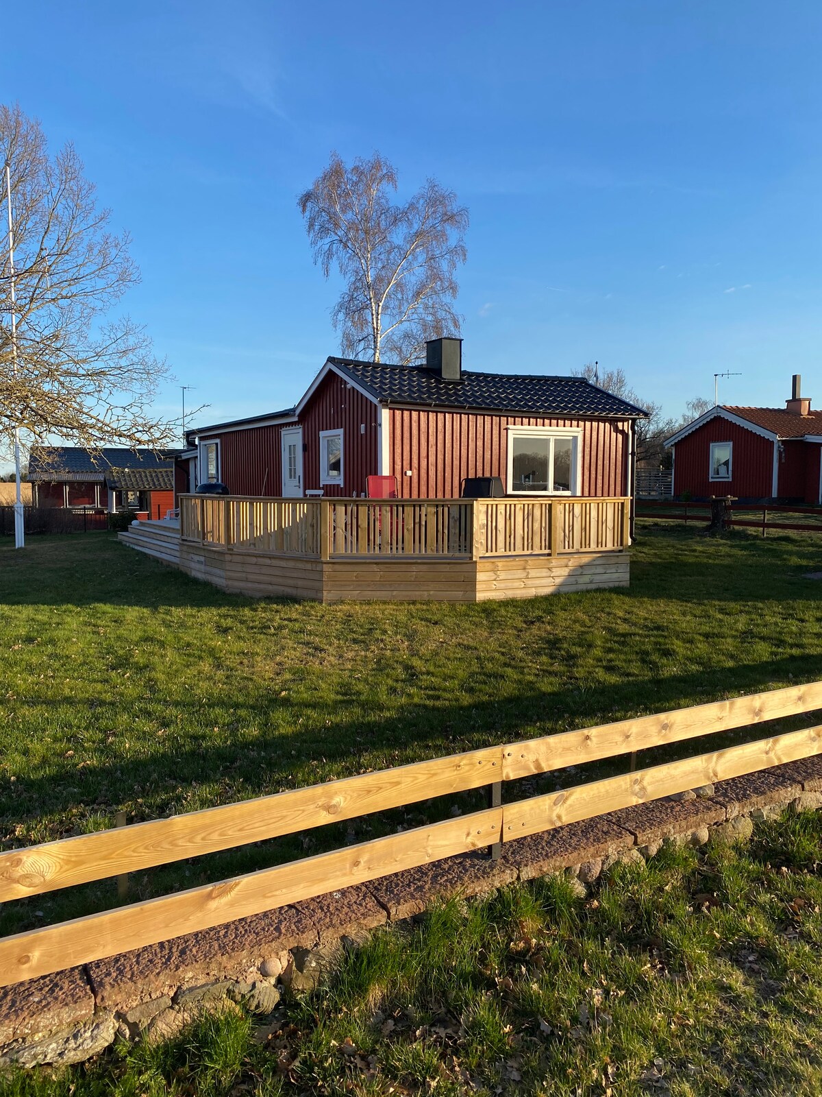 A charming red cottage is showcased, surrounded by a spacious wooden deck. The exterior features a black roof and white-framed windows. Lush green grass surrounds the cottage, with a wooden fence visible in the foreground, complementing the rural setting.