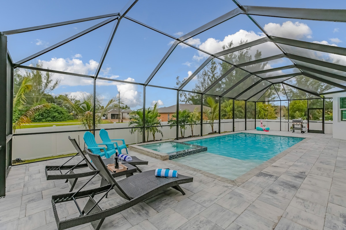 An outdoor space features a screened-in pool area with a rectangular swimming pool and a hot tub. Poolside lounge chairs with blue cushions are arranged around a small table. Lush greenery is visible in the background, enhancing the private ambiance.