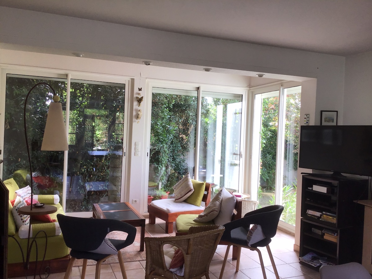 A bright living area is presented with large glass windows that open to a lush garden. Comfortable seating options are arranged, including a mix of chairs and a sofa. A television is situated on a stand, complementing the space with natural light filtering through.