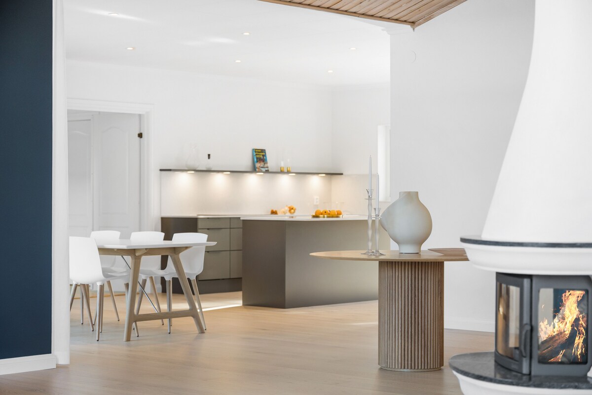 An open-plan living area showcases a modern kitchen with sleek cabinetry and a spacious island. Light wood flooring complements the area, while a cozy fireplace stands nearby, adding warmth to the neutral decor. Simple dining furniture provides a functional and stylish dining space.