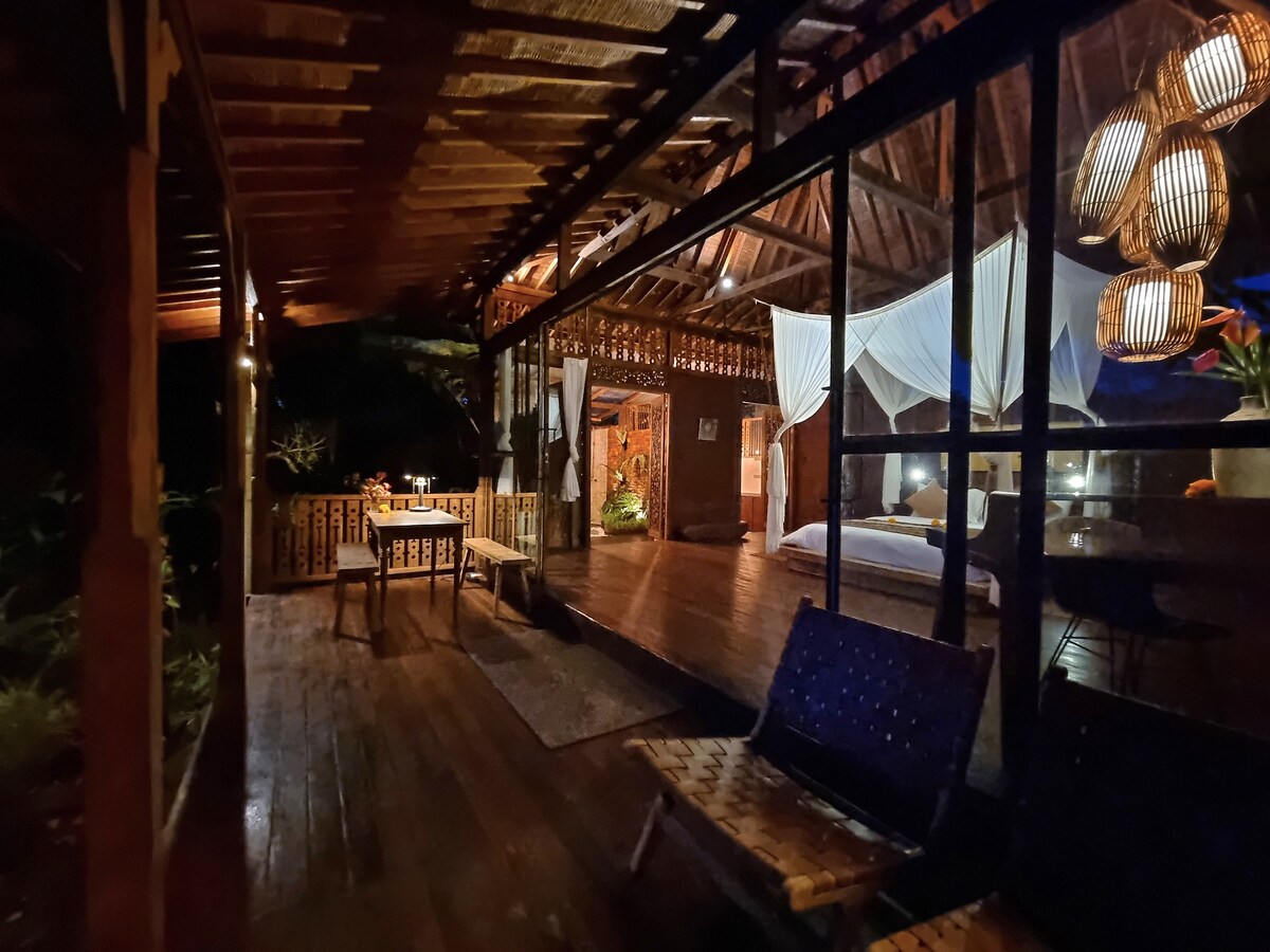A wooden deck area is seen with outdoor seating and a dining table under gently hanging lanterns. Large glass sliding doors lead into the interior space, where a spacious bed is partially visible. Lush greenery surrounds the structure, creating a natural setting.