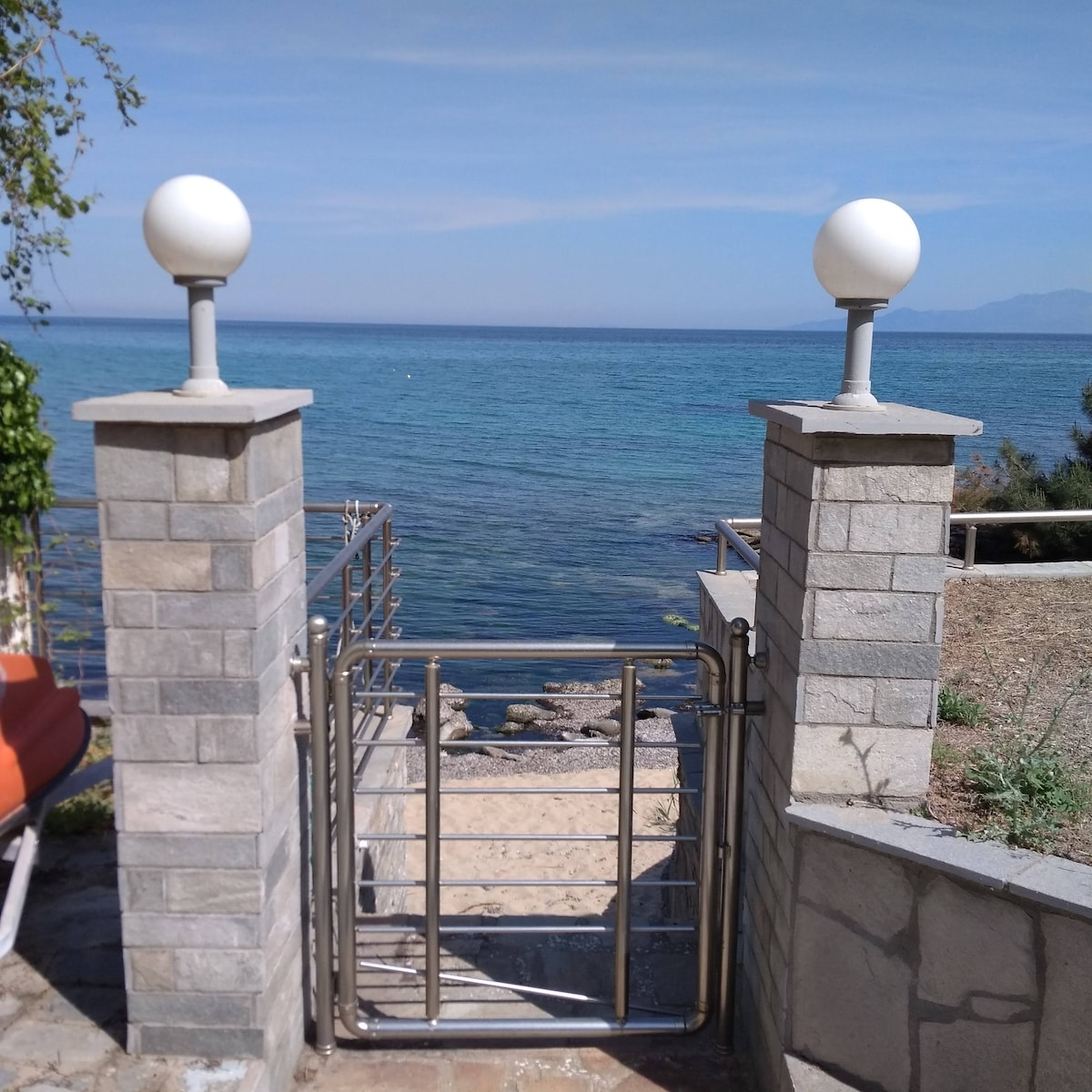 A gated entrance leads to a sandy path towards the sparkling blue sea. Stone pillars frame the gate, topped with round white lights. The calm water stretches into the distance, creating a serene coastal view.