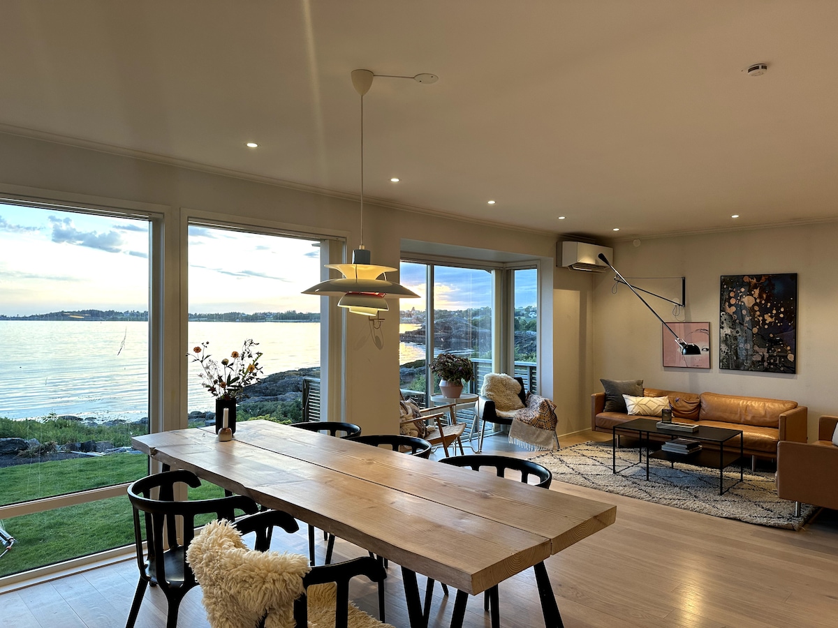 A spacious living area features large windows offering views of the calm water and surrounding landscape. A wooden dining table rests in the center, surrounded by black chairs. A comfortable seating area includes a leather sofa and decorative rugs, creating an inviting atmosphere.