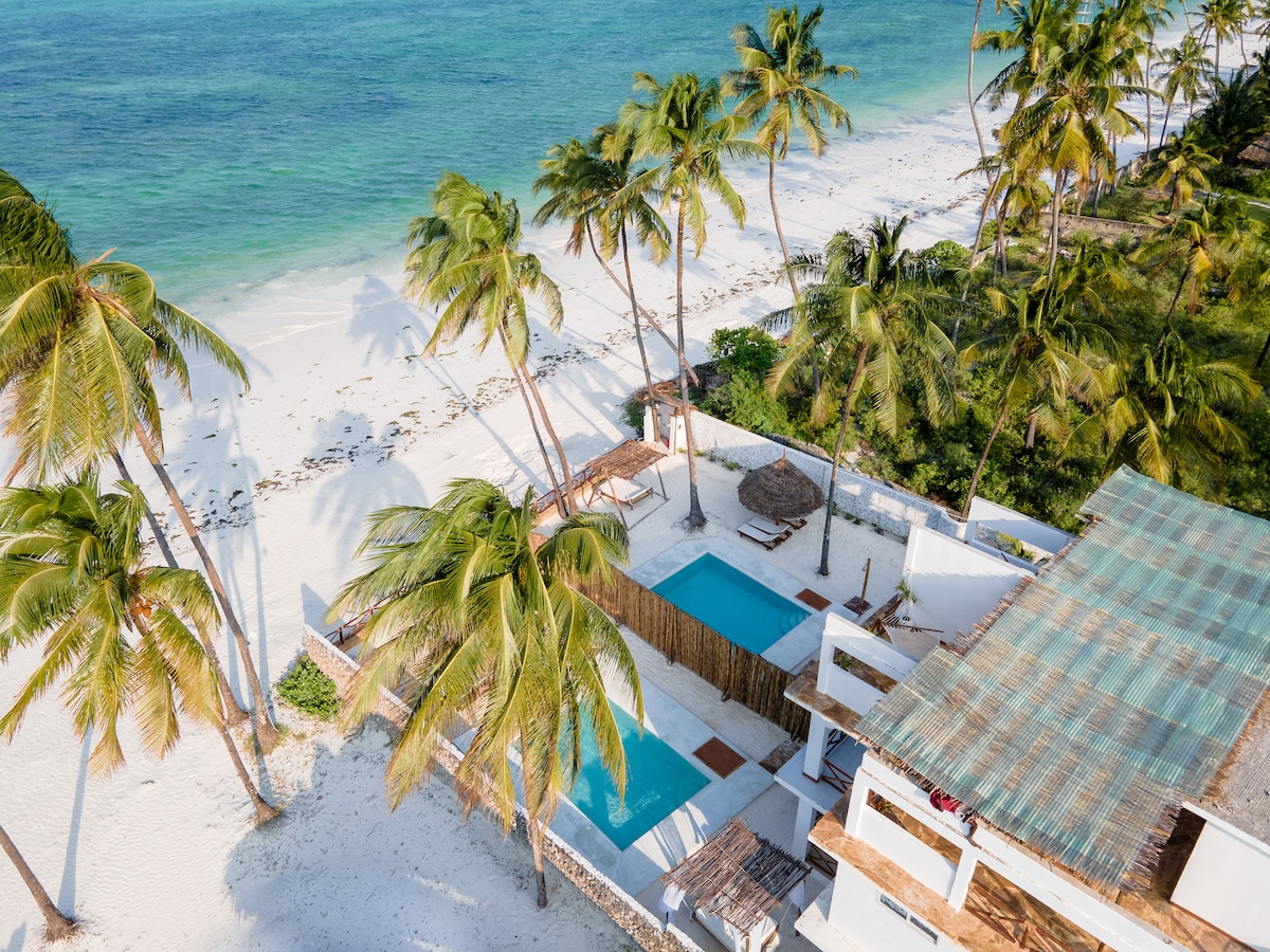 The image captures a serene beachfront setting, featuring a private swimming pool framed by palm trees. The sandy beach extends towards the clear blue waters, with a shaded area and a traditional thatched-roof structure visible nearby.