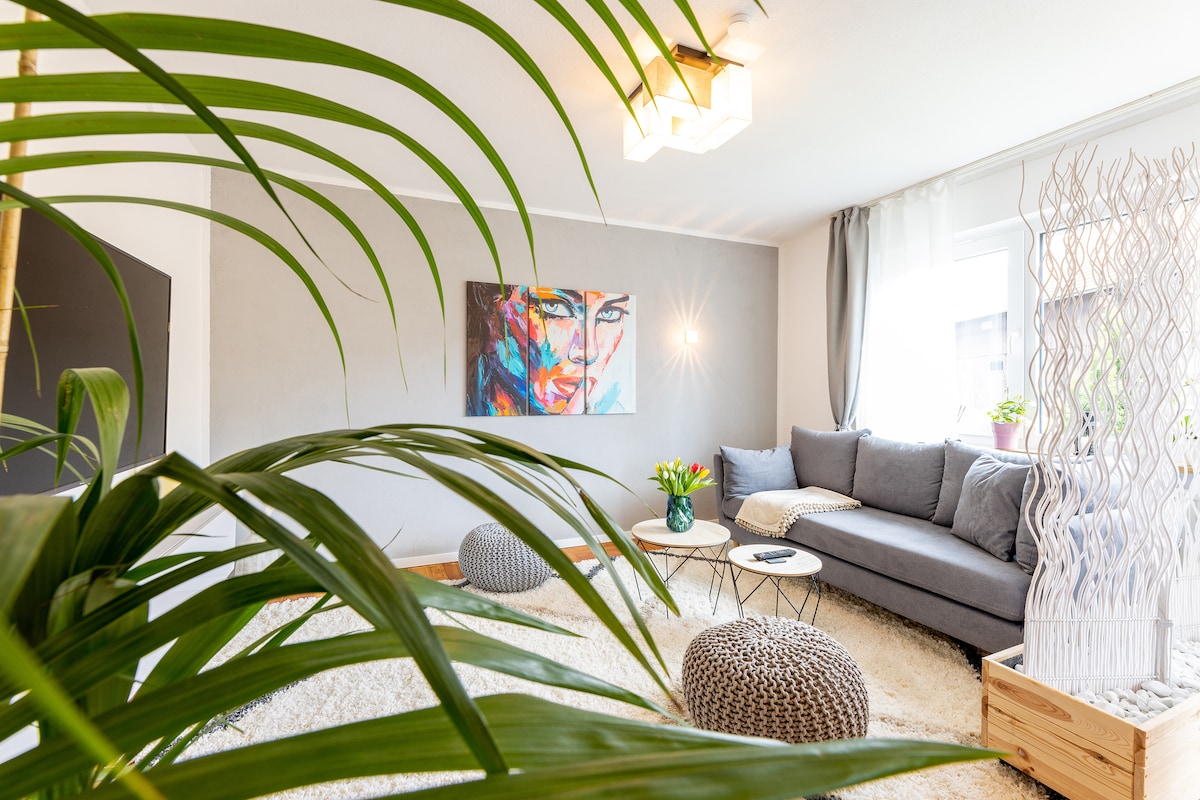 A modern living area is presented with a spacious gray sofa and a round coffee table. A potted plant adds a touch of greenery, while soft light filters through sheer curtains. Decorative elements include a light fixture and artwork on the wall.