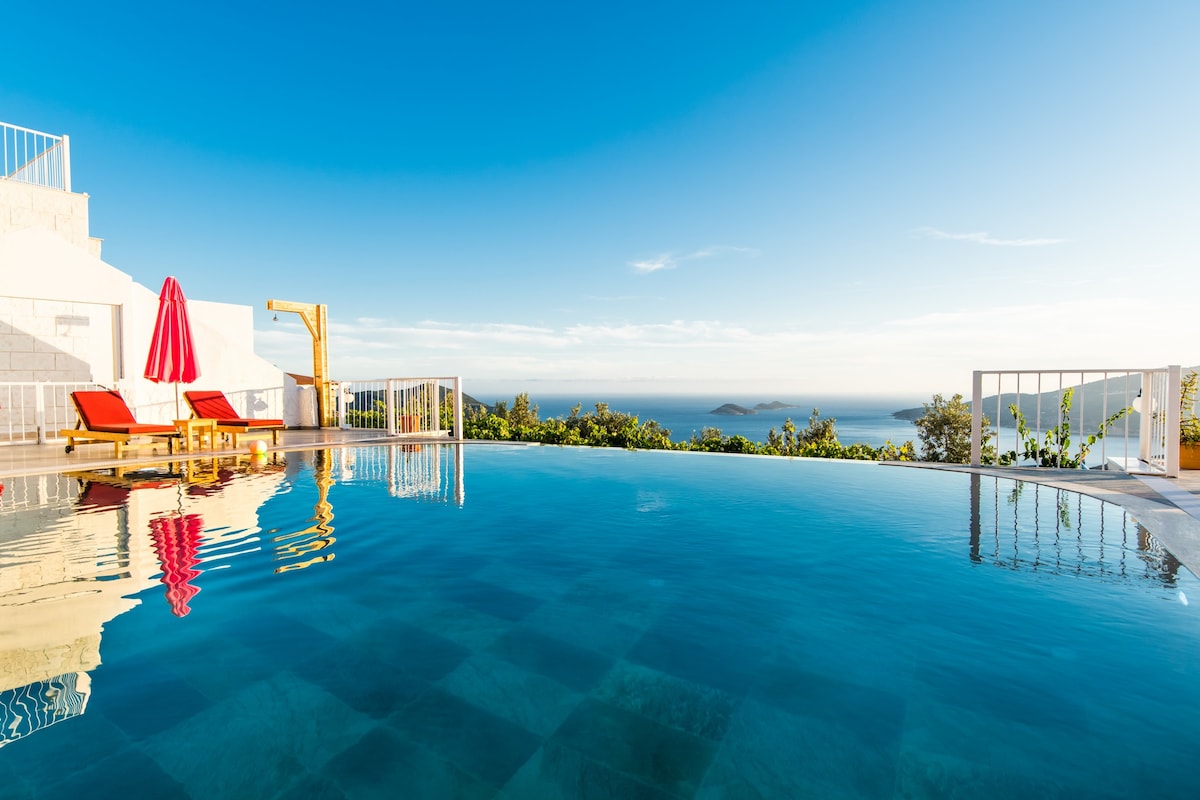 An infinity pool glistens under the clear blue sky, with vibrant red loungers providing seating on the terrace. A red umbrella offers shade, while lush greenery and a panoramic sea view extend beyond the pool's edge.