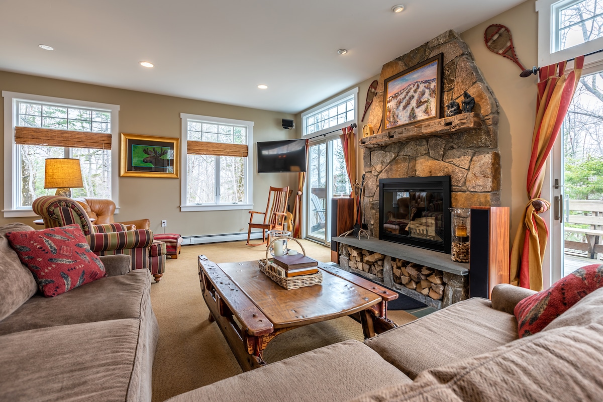 An inviting living area showcases a stone fireplace with a wood log storage beneath. Comfortable seating options include a large sectional sofa and a rocking chair. Bright windows provide natural light and views of the surrounding trees, enhancing the cozy mountain ambiance.