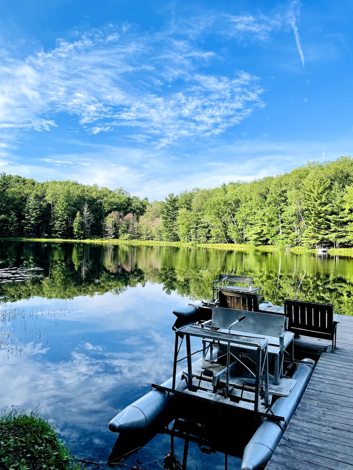 A serene view of Maple Lake is captured, reflecting the clear blue sky and lush green trees. A wooden dock extends into the water, with a small boat and a set of outdoor chairs positioned on it, inviting relaxation and enjoyment of nature.