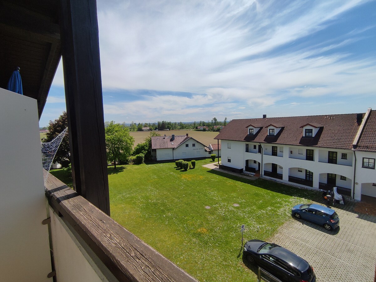 A view from a balcony showcases a spacious green lawn bordered by residential buildings. Several trees provide shade, and distant hills can be seen beyond the flat landscape under a clear blue sky.