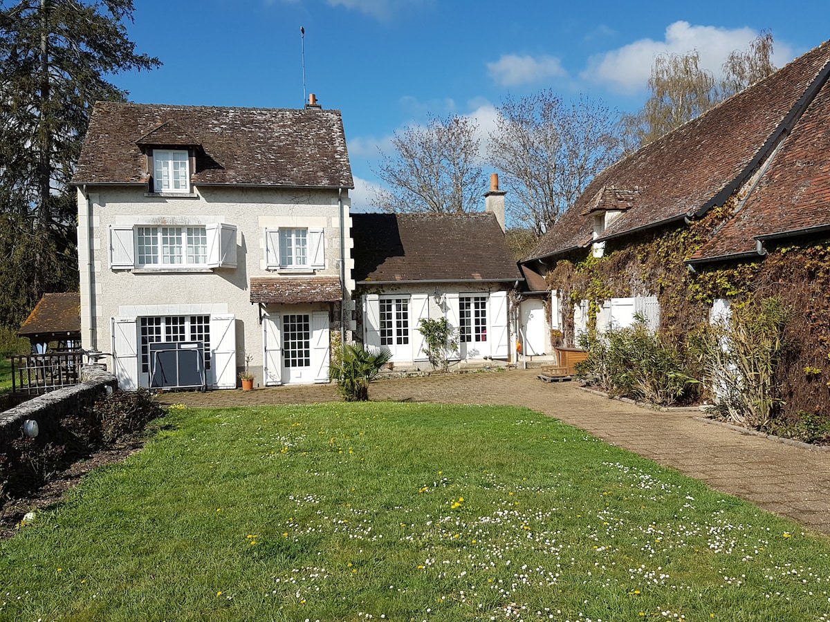 A charming property features a main building with two stories, complemented by an adjacent structure. The spacious front lawn is framed by green grass and dotted with small flowers, while the buildings are accented by a clear blue sky and scattered clouds.