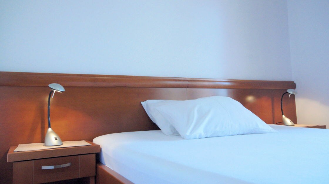 A spacious bed is positioned against a wooden headboard, featuring two bedside tables with sleek lamps on either side. Crisp white linens are arranged on the mattress, and a single white pillow rests at the head of the bed, providing a clean and simple appearance.