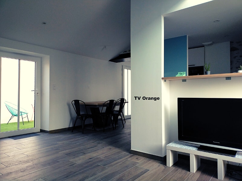 A spacious living area features a dining table with black chairs and a modern television mounted on a simple stand. Light-colored walls and natural tones create a calming environment. A glass door provides access to the outdoors, enhancing the sense of openness.