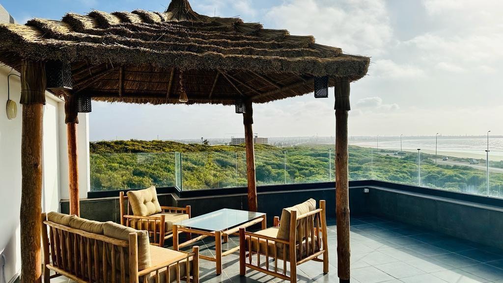 A shaded outdoor seating area features a thatched roof supported by wooden beams. Four wooden chairs surround a glass-topped table, offering a space for relaxation. The backdrop displays a scenic view of lush greenery and the coastline under a cloudy sky.