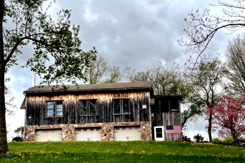 A rustic cabin is depicted, featuring a sturdy wooden exterior with stone accents at the base. Three garage doors are visible below, with an American flag displayed near the doorway. The surrounding landscape includes lush green grass and scattered trees under a cloudy sky.