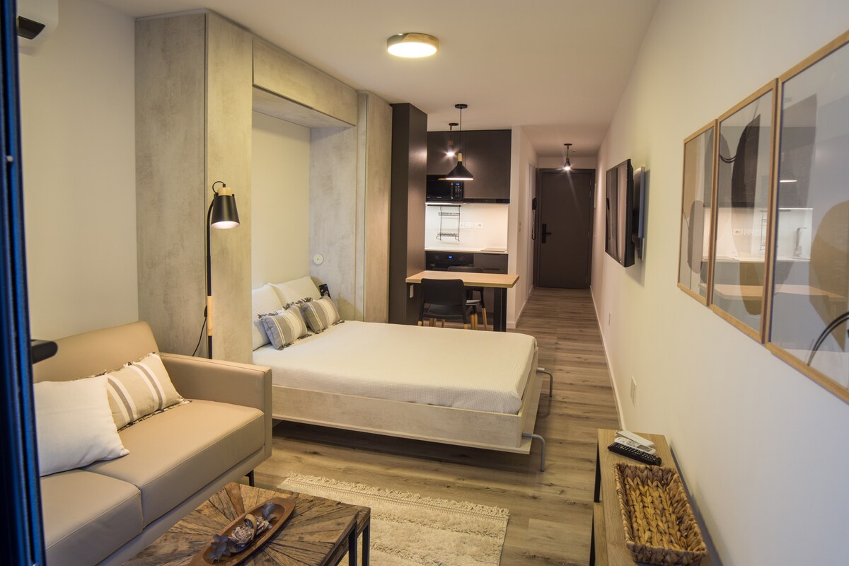 A spacious studio apartment features a foldable bed, modern furniture, and a comfortable sofa. A dining area is present alongside a kitchenette, with light wood flooring and neutral walls creating a serene atmosphere. Natural light enters through large windows, illuminating the functional layout.