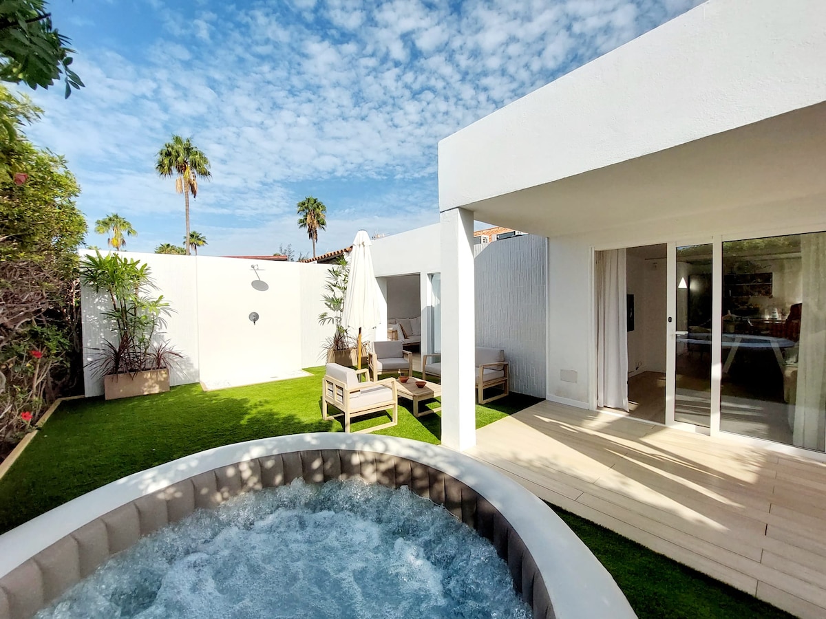 A private garden area features a hot tub surrounded by a neatly trimmed lawn. Comfortable outdoor seating is arranged nearby, while a sun umbrella provides shade. The space is bordered by white walls and lush greenery, creating a serene and inviting atmosphere.