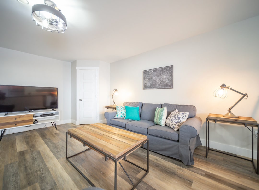 A modern living area features a grey sofa adorned with vibrant teal and patterned cushions. A stylish wooden coffee table is centered in the room, complemented by two industrial lamps on either side. A flat-screen television is mounted on a light wall, enhancing the open space.