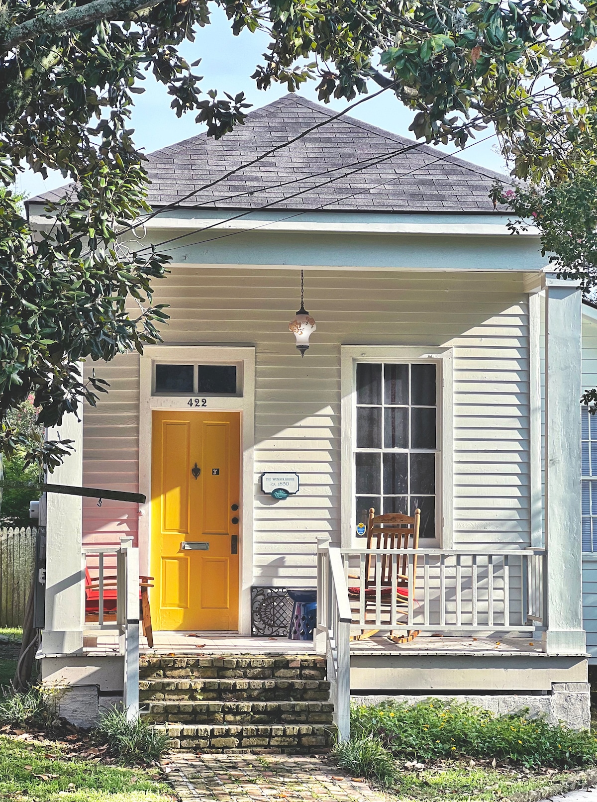 A charming cottage exterior features a vibrant yellow front door that adds a pop of color. Light wood siding is complemented by large windows. A small porch area includes a rocking chair and a nearby lamp, inviting relaxation. Lush greenery surrounds the entrance.