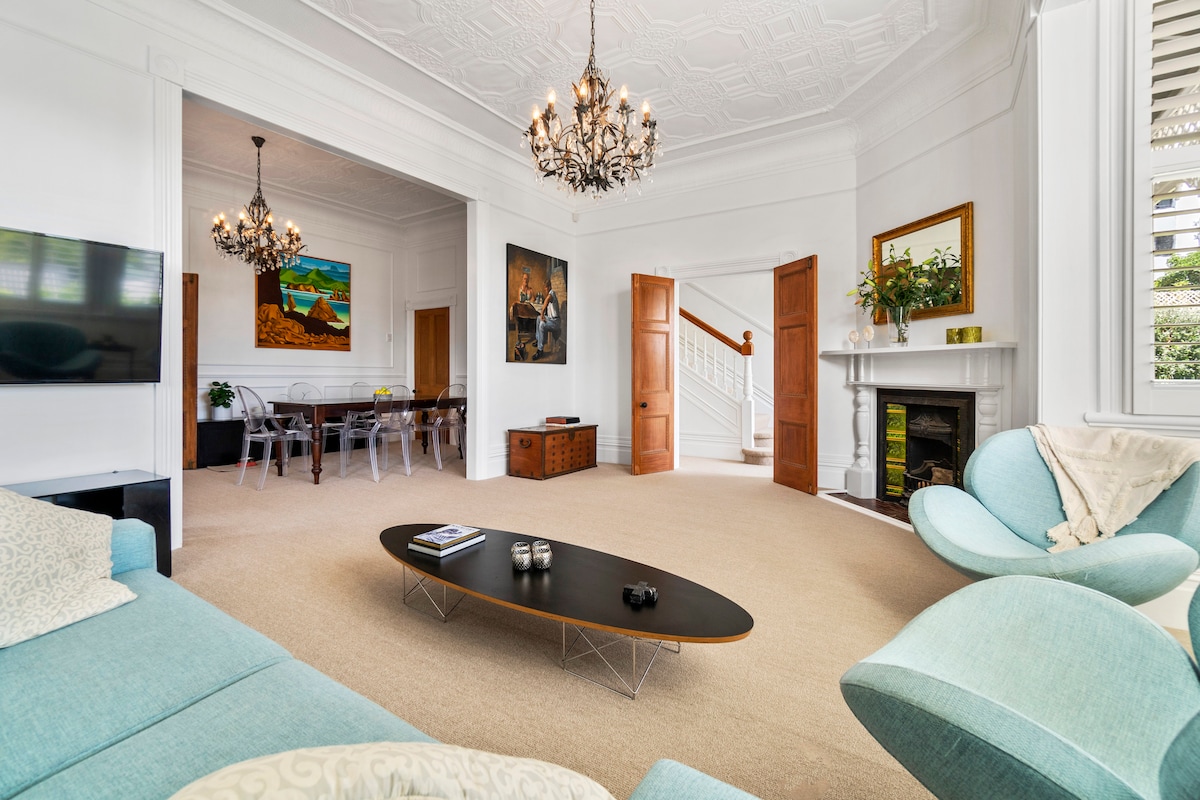 A spacious living area is highlighted by soft teal furniture and an elegant coffee table with a smooth surface. Large windows allow natural light to fill the room, enhancing the bright and open atmosphere. A tasteful chandelier hangs from the intricately designed ceiling.