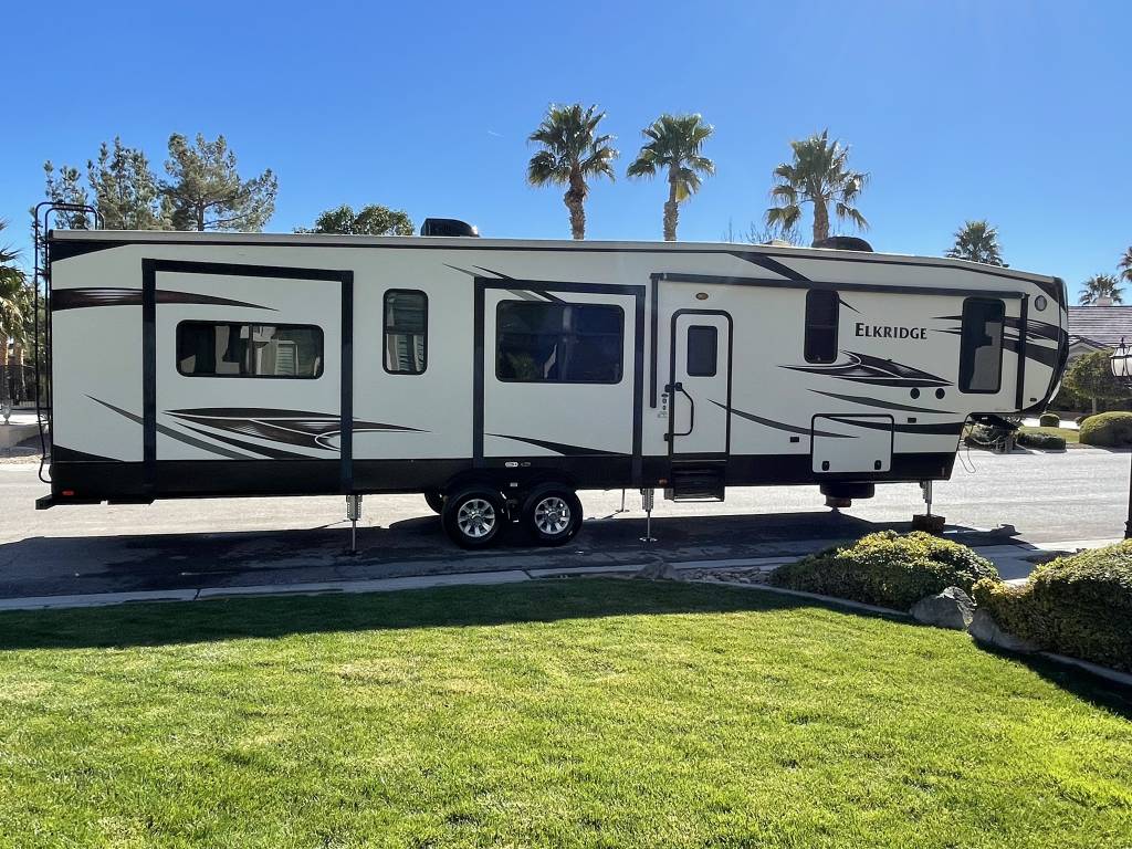 A modern RV is showcased, parked on a green lawn with neatly trimmed grass. Palm trees can be seen in the background, adding a serene touch to the setting. The RV features multiple windows and a sleek exterior design.