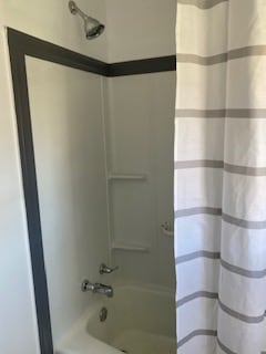 The bathroom features a shower area enclosed by a white shower curtain with gray stripes. Shower fixtures, including a handheld showerhead and a tub spout, are installed on the wall. The walls are painted in a light color, creating a clean and simple design.