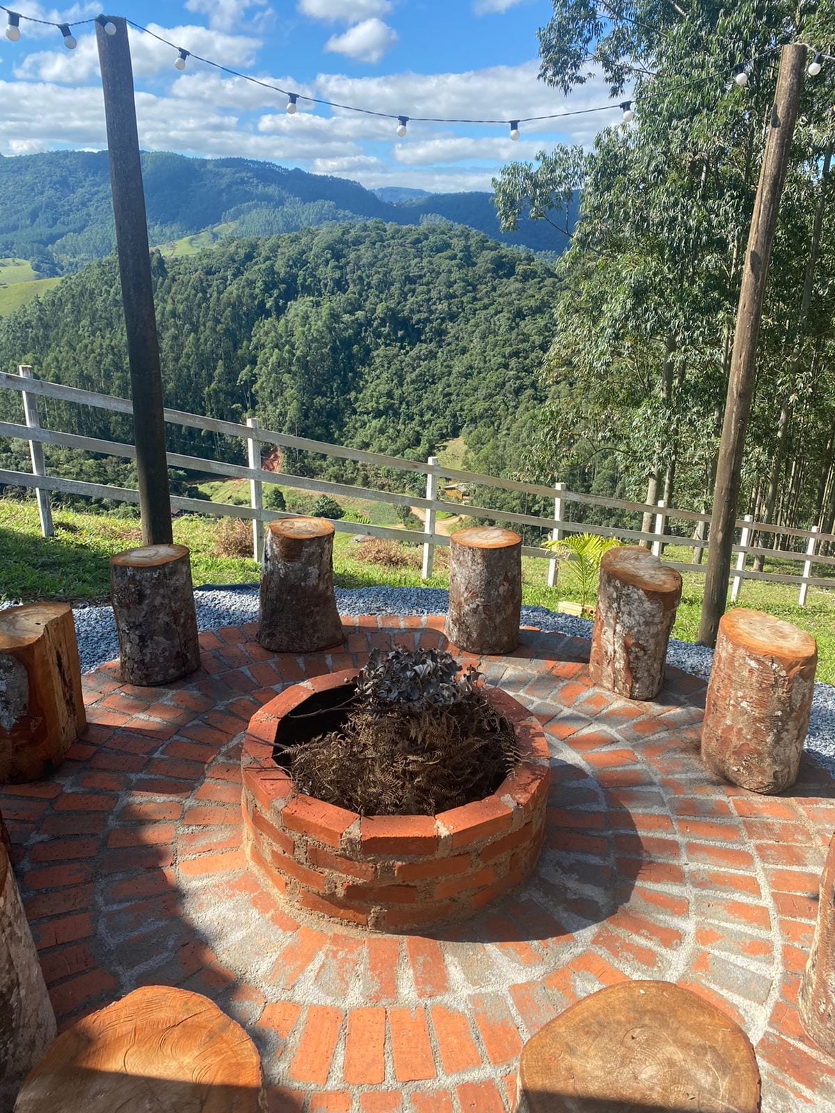 A circular fire pit is surrounded by rustic log seating, positioned on a terrace overlooking a lush mountainous landscape. The sky is dotted with clouds, and string lights are visible overhead, enhancing the cozy outdoor gathering space.