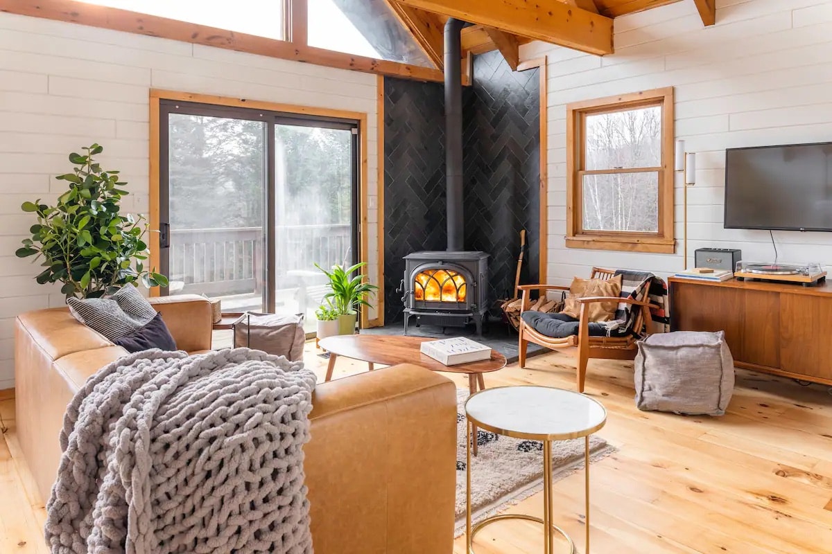 A cozy living area is highlighted by large windows allowing natural light to fill the space. A wood-burning stove serves as a focal point, with a plush sofa draped in a soft throw. A mix of plants and decorative items enhances the inviting atmosphere.