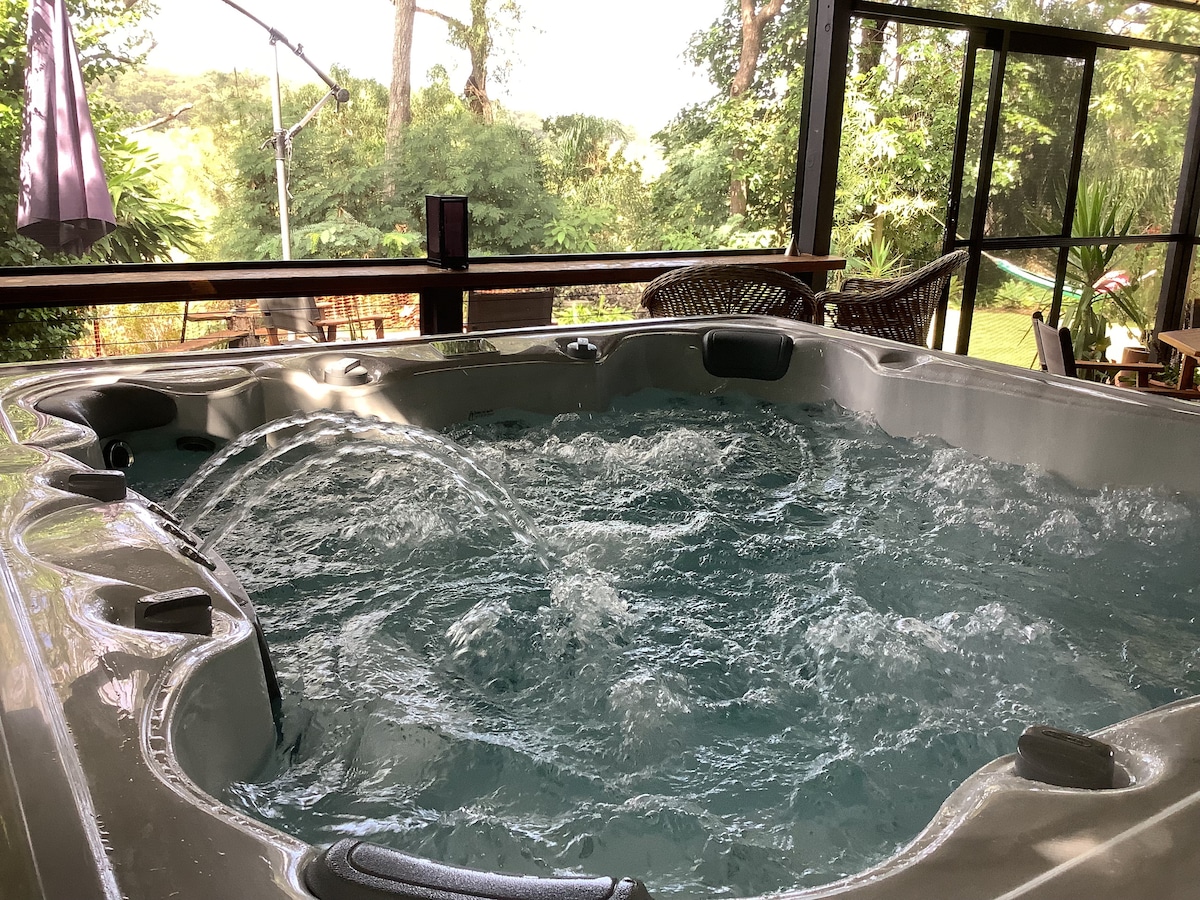 A hot tub with bubbling water is situated indoors, surrounded by windows that offer views of lush greenery. Natural light enters the space, highlighting the sleek design of the hot tub and creating a fresh ambiance throughout the area.