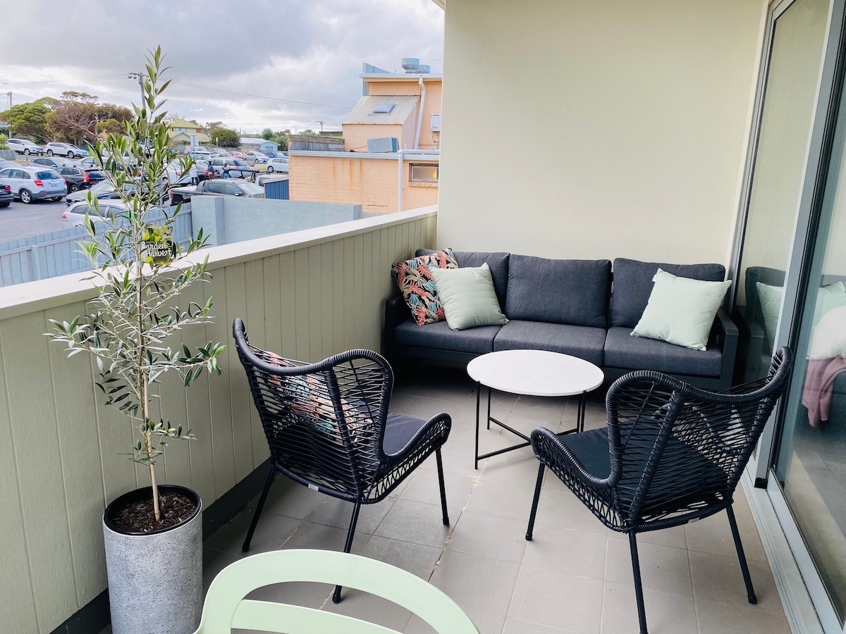 A balcony space is presented with modern wicker chairs and a circular table. A sleek couch adorned with pillows offers comfortable seating. A potted olive tree adds a touch of greenery, while nearby expansive windows provide ample natural light.