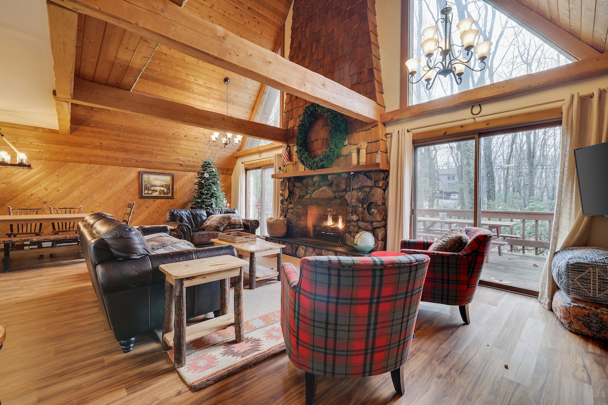 A spacious living area features a vaulted wooden ceiling with exposed beams. A stone fireplace serves as a focal point, surrounded by comfortable seating, including a leather sofa and plaid chairs. Large windows offer a view of the wooded outdoors, with soft natural light illuminating the room.