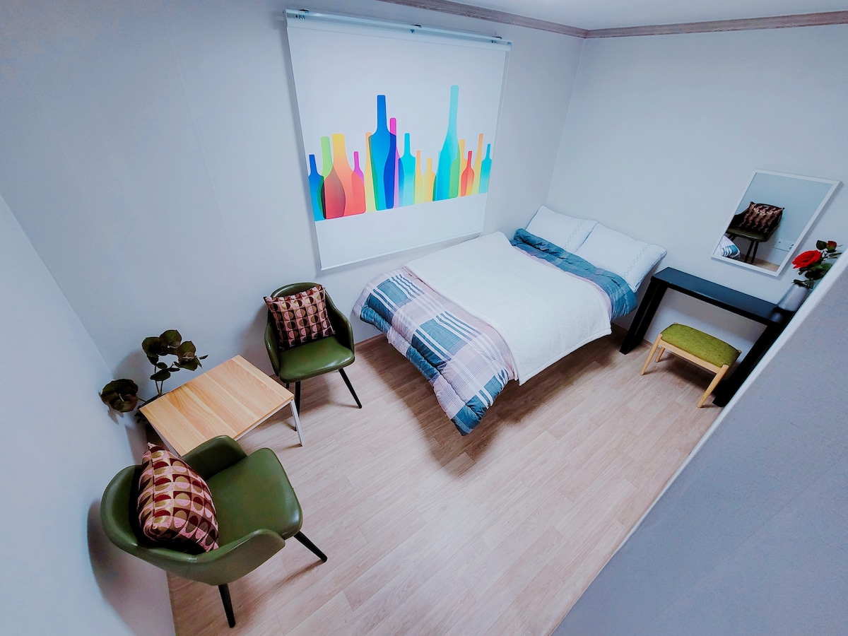 A cozy sleeping area is presented, featuring a spacious bed with a light blanket layered over it. A colorful wall art piece depicting bottles adds a touch of vibrancy. A small wooden table and two chairs, one green and one with patterned upholstery, are arranged nearby.