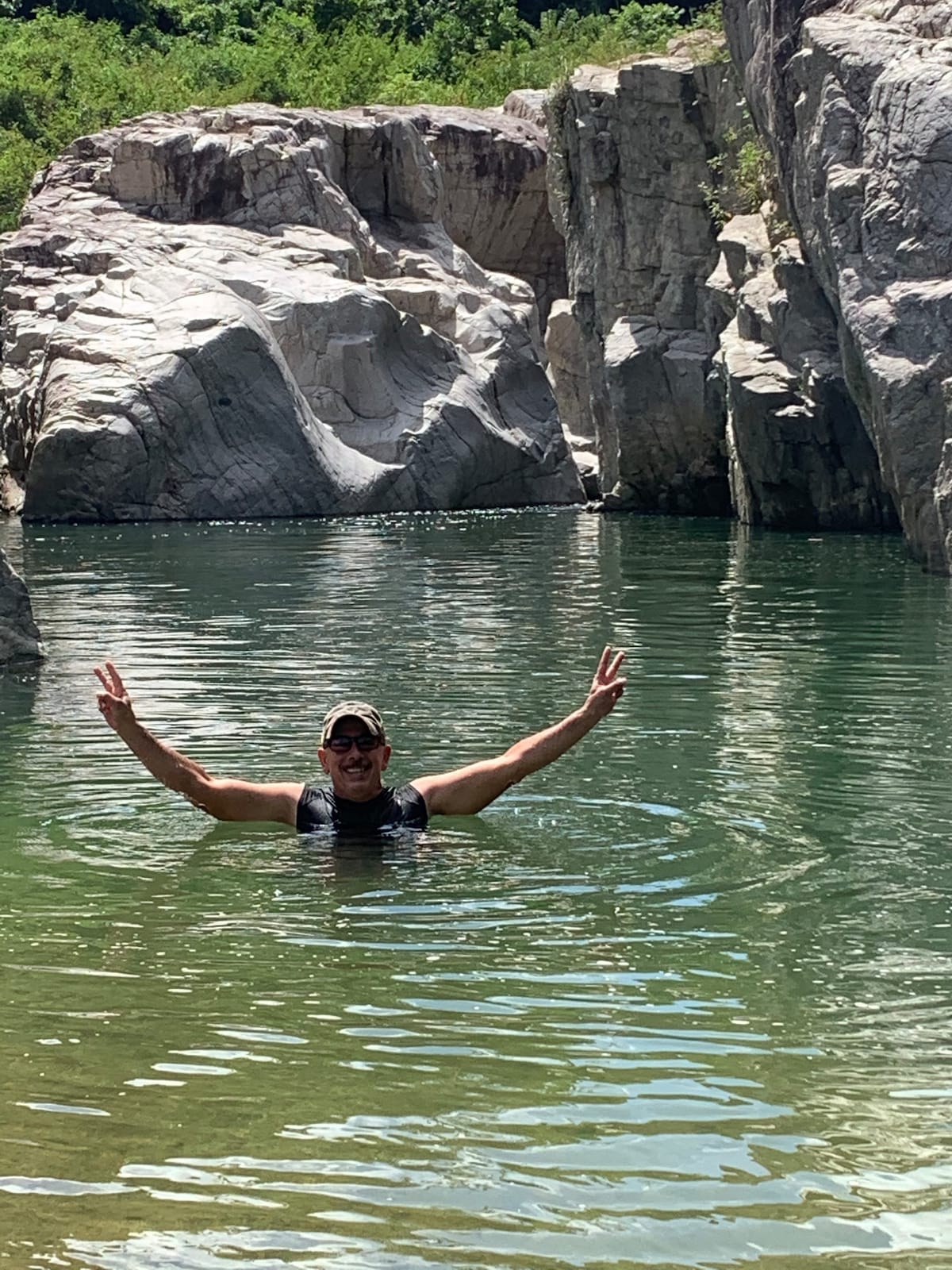 A person stands in a tranquil body of water, surrounded by tall, rugged rock formations. The water appears clear and inviting, with gentle ripples reflecting the natural landscape. Lush greenery is visible in the background, enhancing the serene atmosphere of this secluded area.