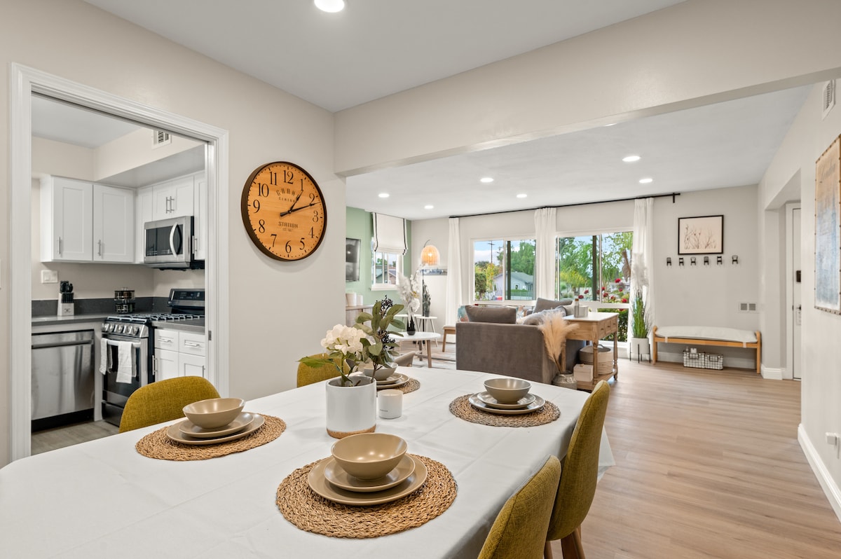A dining area is highlighted by a neutral-toned table set with plates and woven placemats. A large wall clock adds character. The space seamlessly connects to a modern kitchen with stainless steel appliances, and a living area can be seen in the background, featuring natural light and comfortable seating.