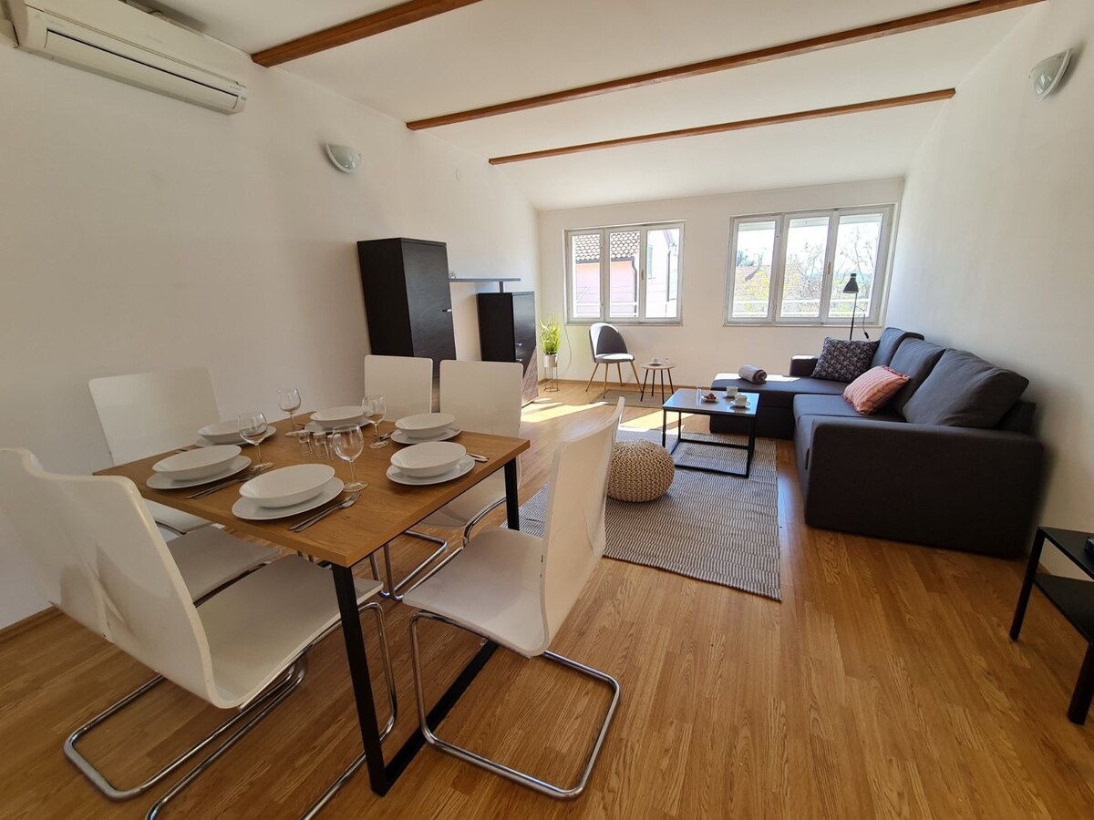 A bright living area features large windows that invite natural light, highlighting a comfortable sofa and a dining table set for six. The space has a simple, modern design with wooden floors and minimalistic decor, creating an open and airy atmosphere.