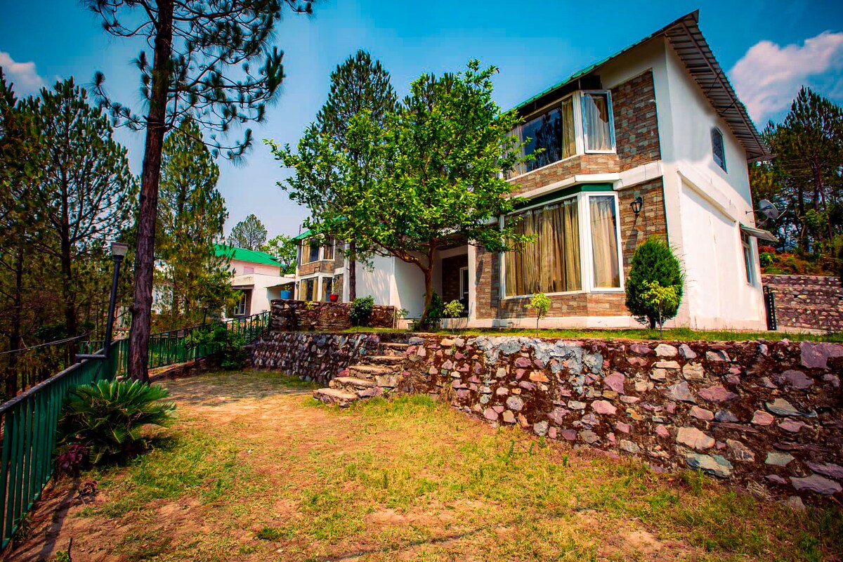 A two-story villa is surrounded by lush greenery and towering pines. Large windows reflect natural light, while a stone pathway leads up to the entrance. Carefully tended shrubs and a small garden enhance the inviting exterior, creating a serene environment.