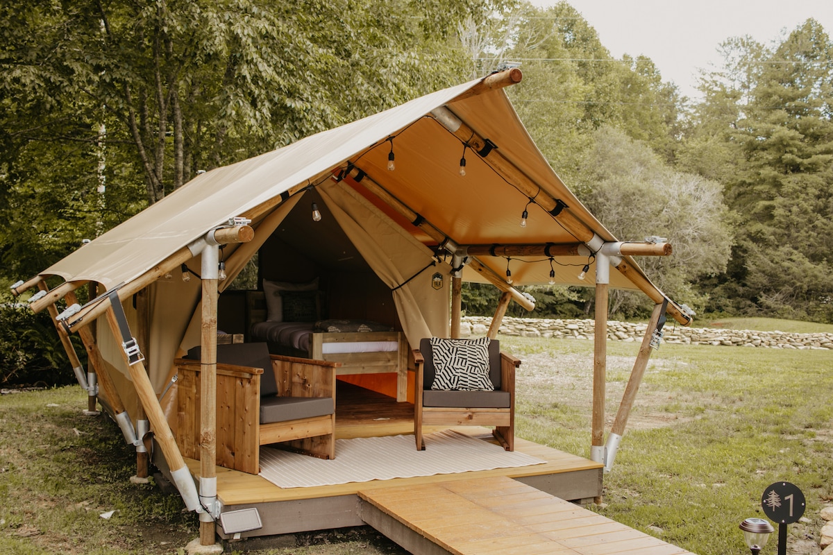 A safari tent is set on a grassy area, featuring a wooden porch with steps leading to a cozy seating area. Two twin beds are visible inside, surrounded by natural greenery and strings of lights hanging from the tent’s structure.