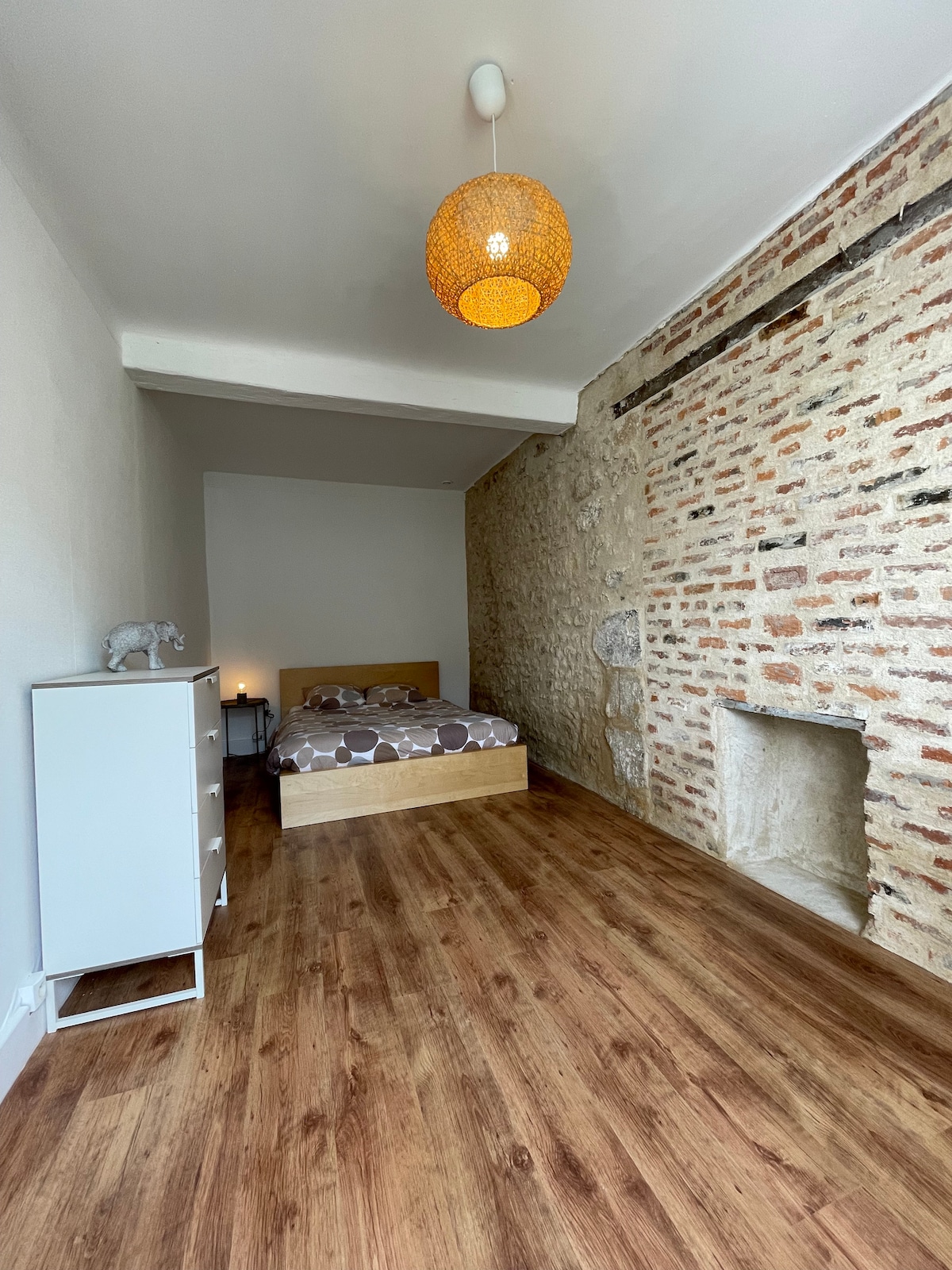 A spacious bedroom features a wooden bed with neutral bedding. An accent wall of exposed brick adds character, while a simple dresser stands nearby. The warm glow of a pendant light illuminates the room, creating a calming environment. A decorative element can be seen on the bedside.