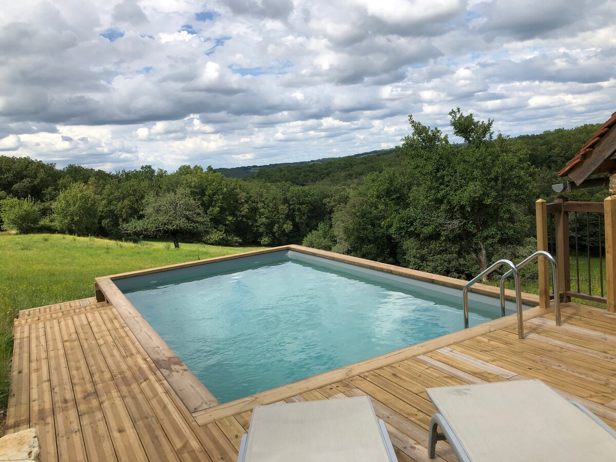 An outdoor wooden deck surrounds a rectangular pool, reflecting the sky's clouds. Lush greenery and distant hills provide a natural backdrop. Two lounge chairs are positioned nearby, offering a space for relaxation.