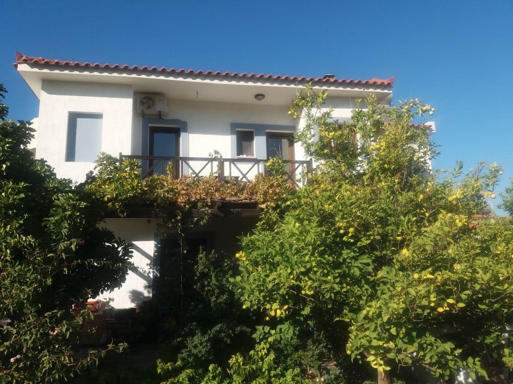 The exterior of a two-story house is framed by lush greenery and vibrant vines. The balcony extends from the upper floor, providing a view of the garden below. Large windows reflect natural light, and the clear blue sky complements the tranquil setting.