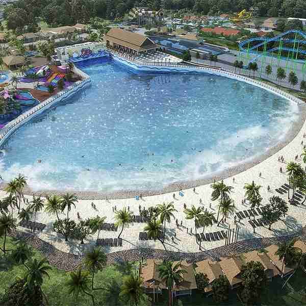 An aerial view of a large wave pool is presented, surrounded by sandy beach areas. Palm trees are visible along the perimeter, and colorful attractions can be seen in the background, with lounge chairs arranged on the beach for guests. The clear blue water creates a vibrant atmosphere.
