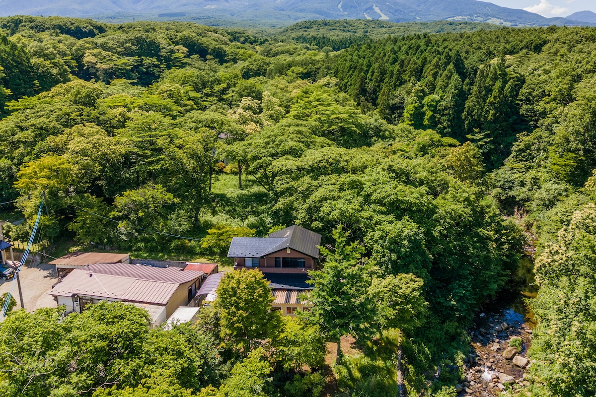 Aerial view of the property surrounded by lush greenery, with a small creek flowing beside it. The building layout includes a mix of traditional and modern architecture, harmonizing with the natural landscape and providing a serene retreat atmosphere.