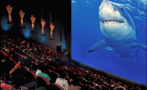 A full cinema view is captured, featuring a large screen displaying a shark in an ocean scene. In front, numerous audience members are seated in plush red chairs, engaged in the film, with the theater's elegant wall sconces softly illuminating the space.