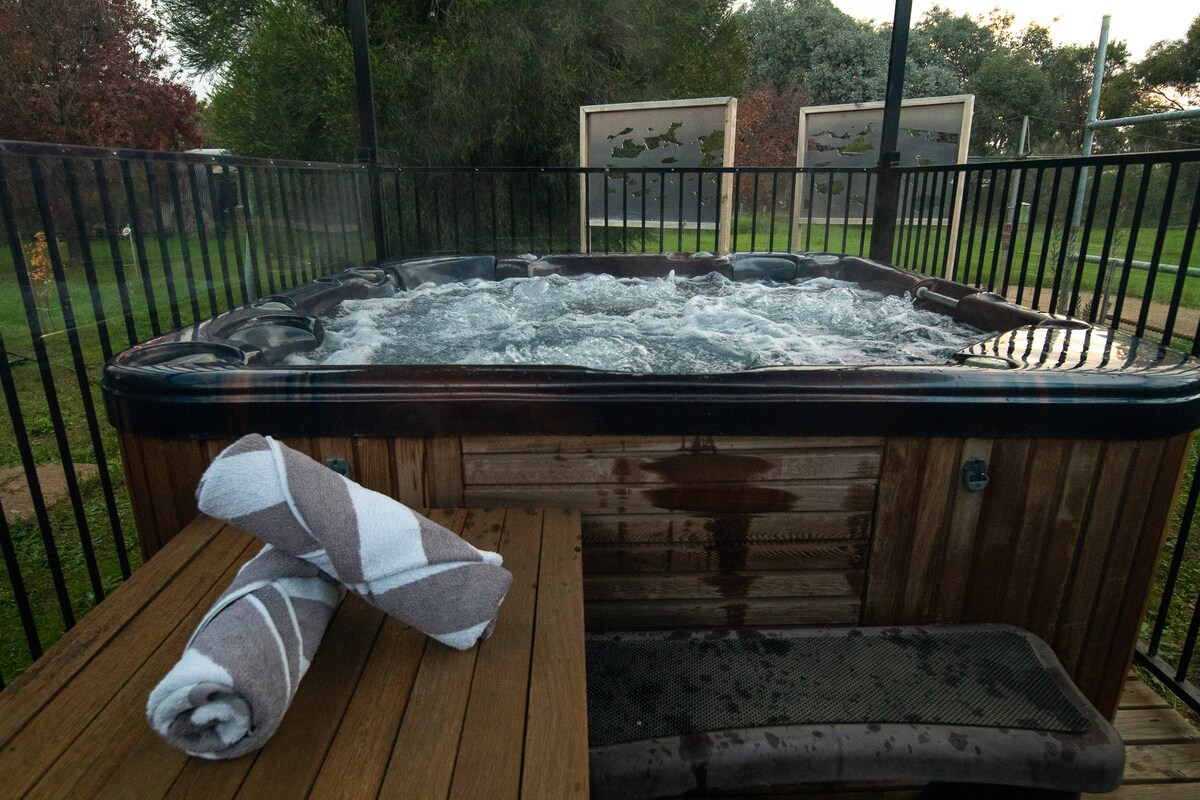 A hot tub is featured on a wooden deck, surrounded by a black fence for privacy. Two rolled towels are neatly placed on the edge, with bubbles and steam visible on the surface of the water, creating an inviting space for relaxation.