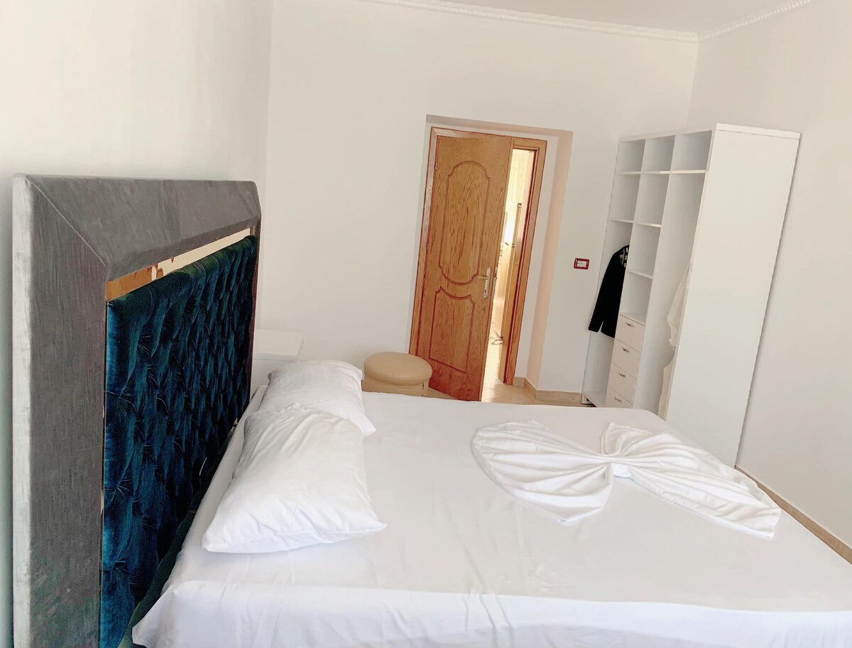 A well-appointed bedroom features a spacious bed with white linens and decorative pillows. A stylish headboard adds a modern touch. In the background, a wooden door opens to a hallway, and a white wardrobe stands against the wall, providing additional storage.