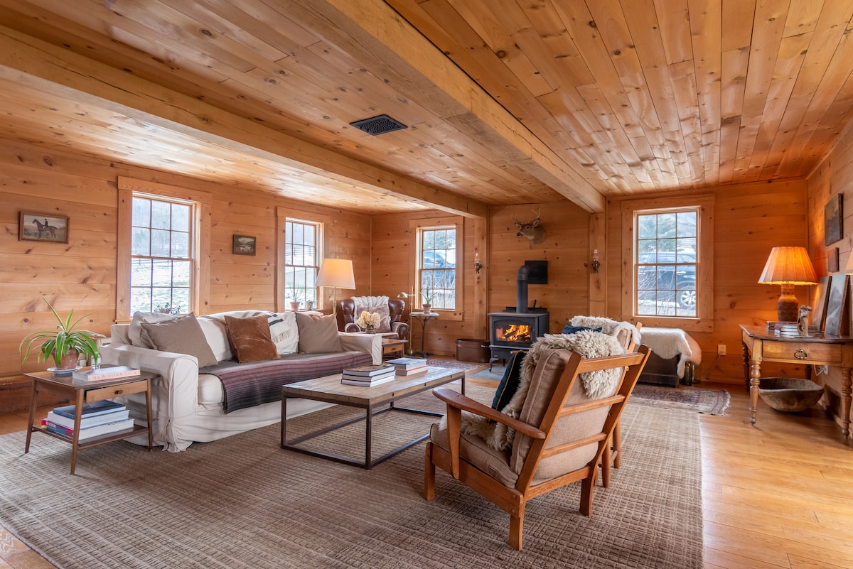 A spacious living area features wooden walls and a ceiling with exposed beams. Natural light illuminates the room through multiple windows. A sofa and two armchairs are arranged around a coffee table, with a small wood stove providing warmth.